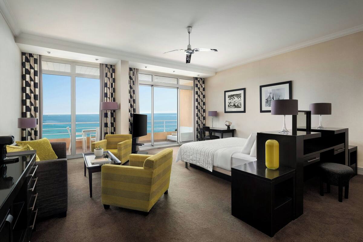 A hotel room with a bed, a desk, a chair, a sofa, and a TV. There are large windows with a view of the ocean.