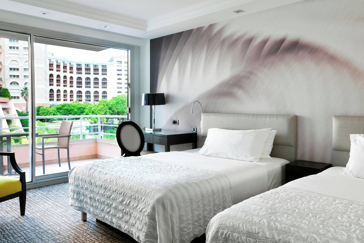 A hotel room with two beds, a large window with a view of buildings and greenery, a chair, and a desk with a lamp.