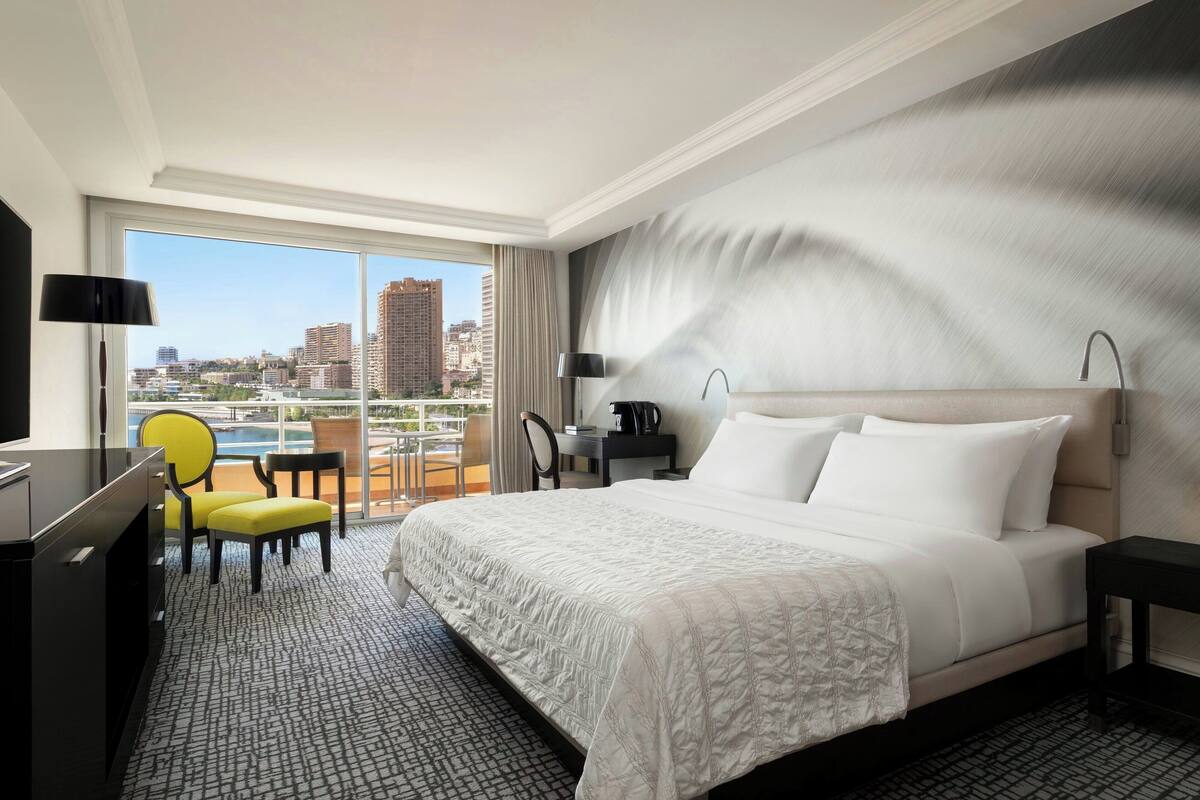 A hotel room with a large bed, a desk, a chair, a view of the city, and a balcony.