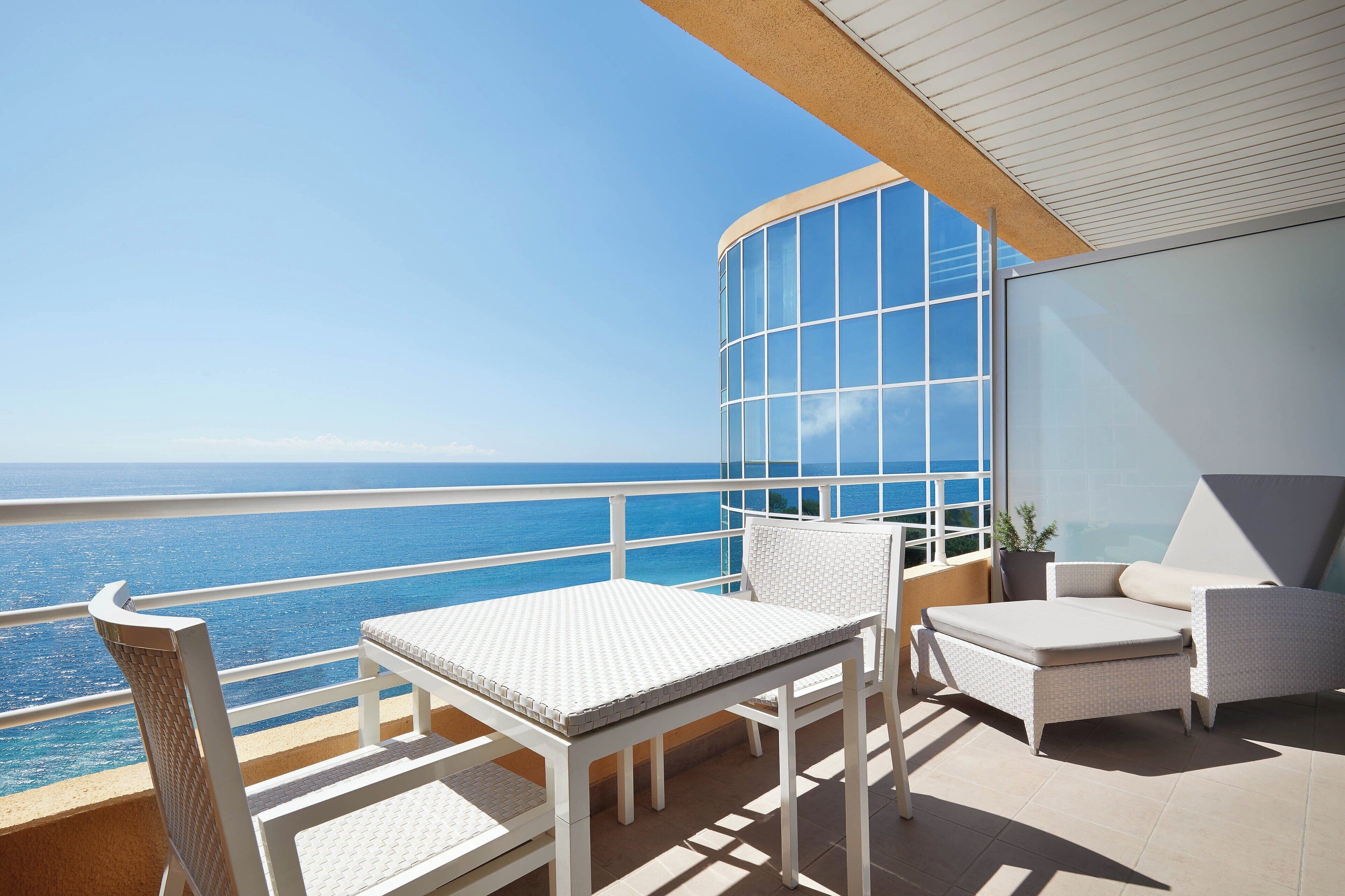 A balcony with a white table and chairs, a potted plant, and a view of the ocean.