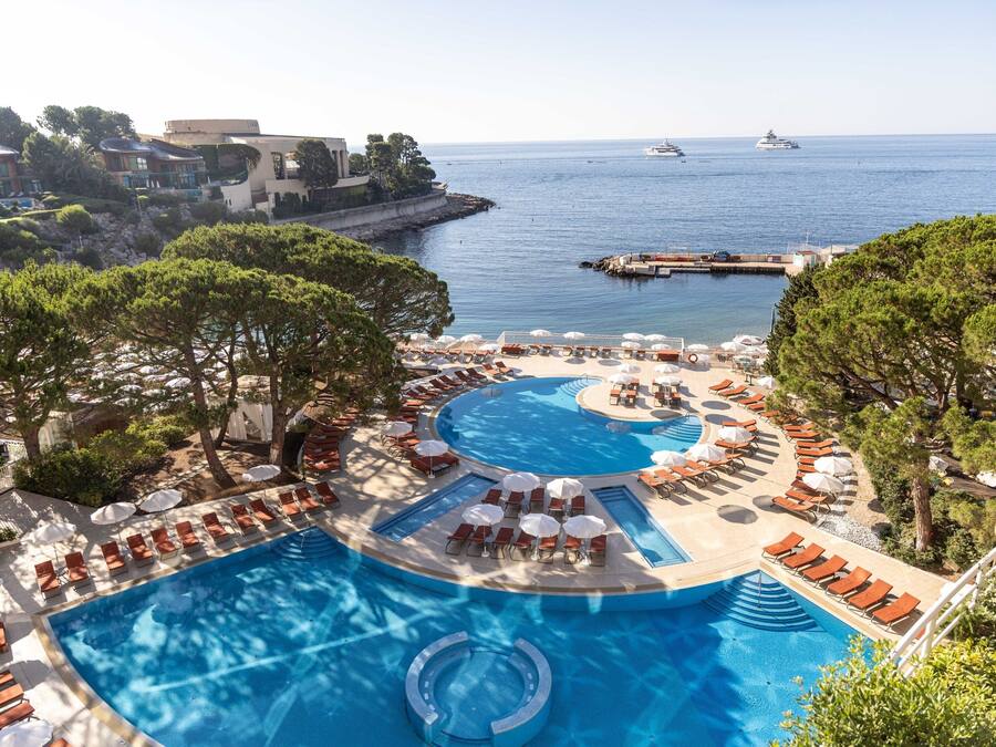 A resort pool with multiple sunbathing areas, a view of the sea, and a dock with boats.