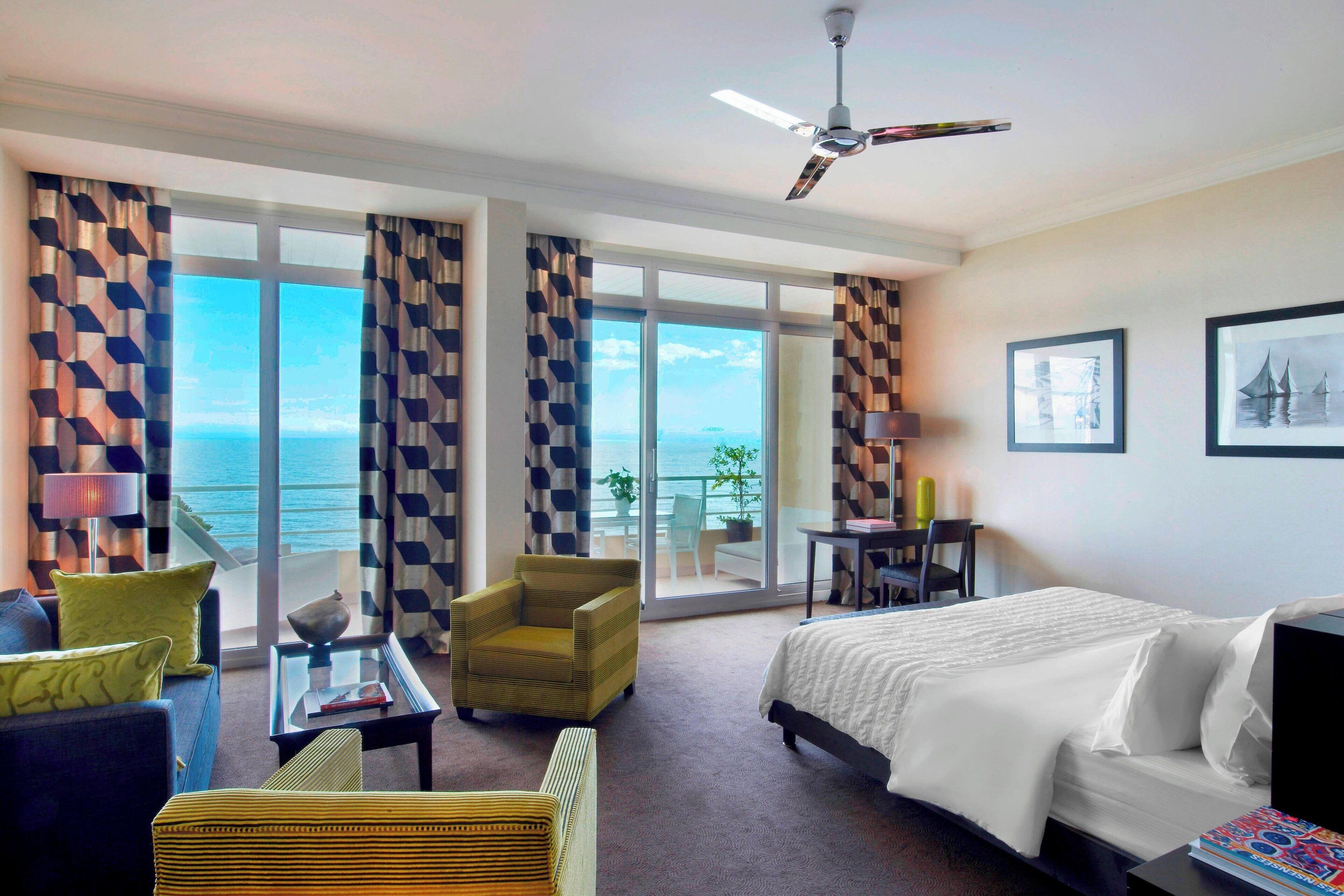 A hotel room with a bed, a sofa, a chair, a table, and a balcony with a view of the sea.