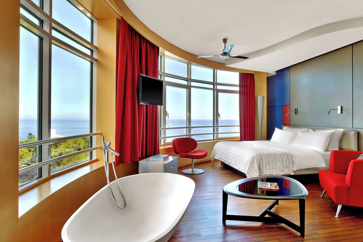 A modern hotel room with a large window, a bathtub, a bed, a red chair, and a television.