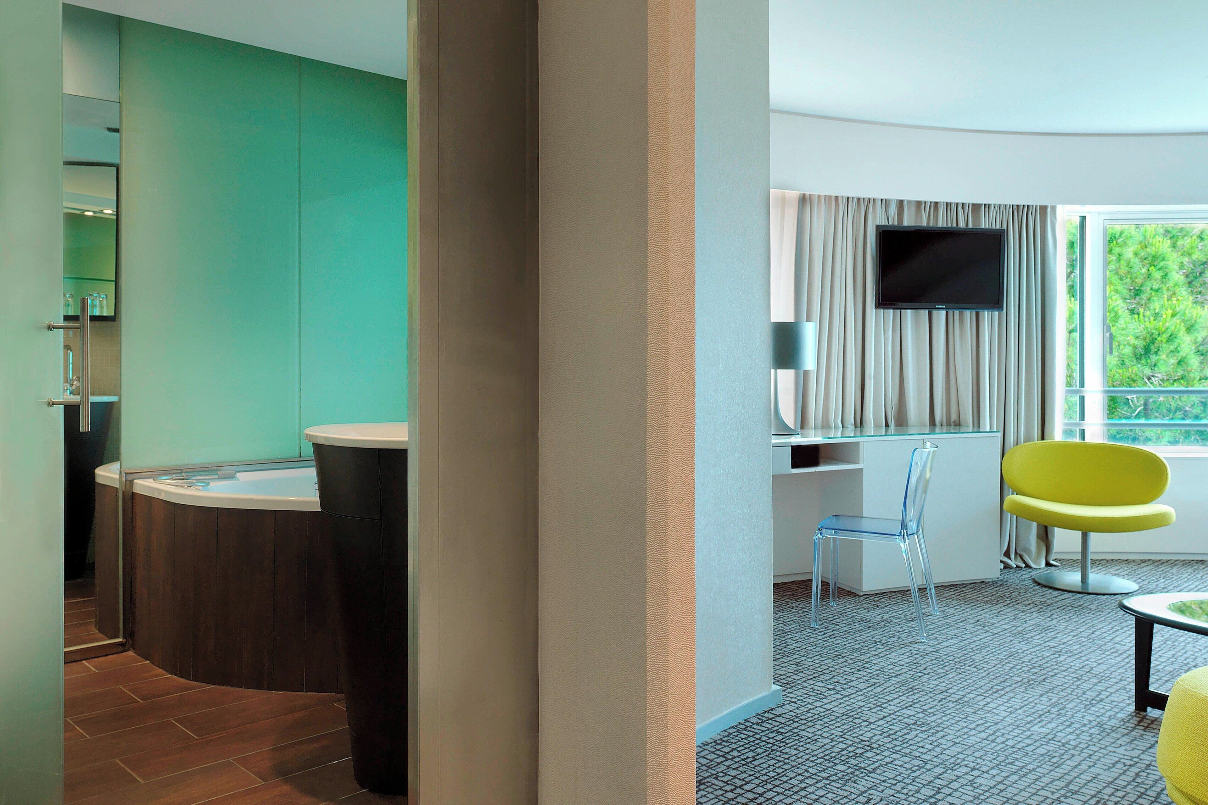 Une chambre d’hôtel moderne avec une baignoire à formes arrondies, un téléviseur à écran plat, un fauteuil jaune et une vue sur la verdure.
