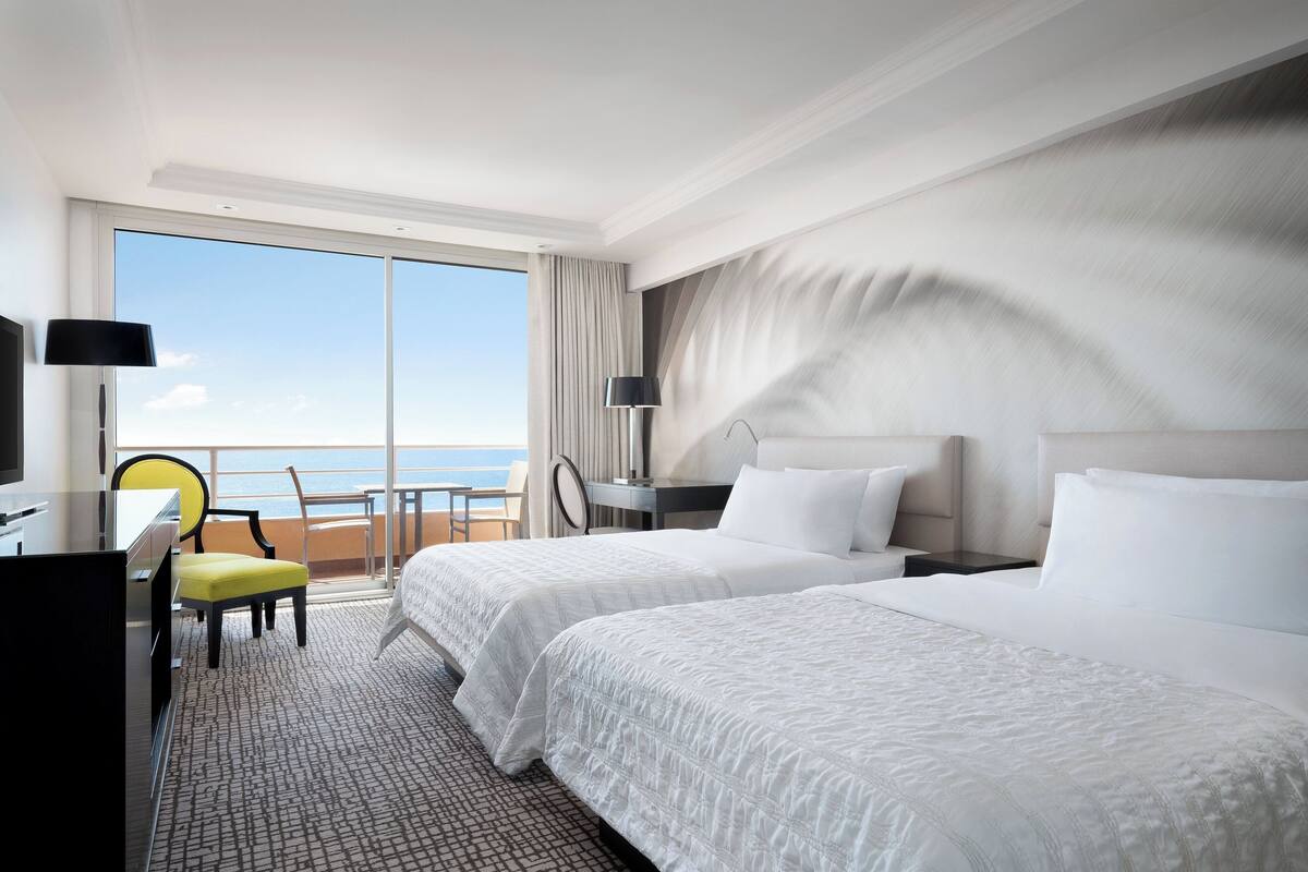 A hotel room with two beds, a desk, a chair, and a view of the sea.