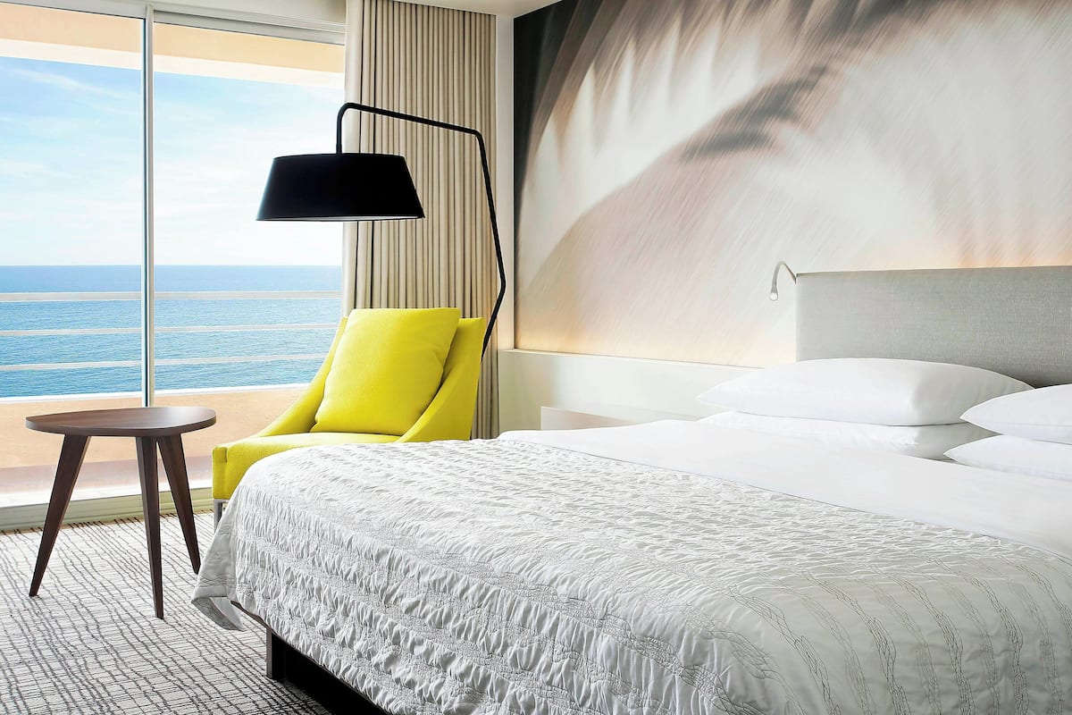 A hotel room with a large bed, a bright yellow armchair, a small round table, and a view of the ocean through large windows.
