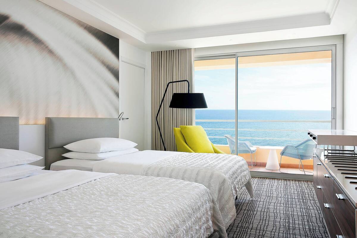 A hotel room with two beds, a large window with a view of the sea, a floor lamp, and a small table with chairs.