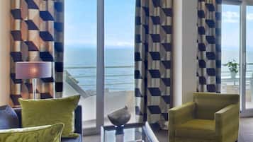 A modern living room with a view of the sea, featuring geometric patterned curtains, a sofa, two armchairs, and a coffee table.