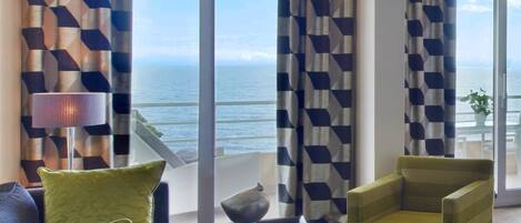 A modern living room with a view of the sea, featuring geometric patterned curtains, a sofa, two armchairs, and a coffee table.
