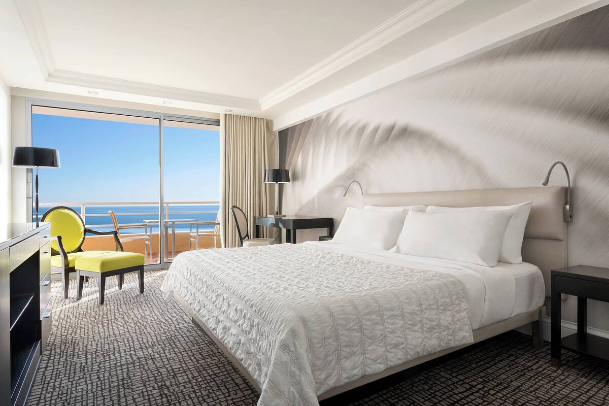 A hotel room with a large bed, a desk, a chair, a TV, and a view of the ocean.