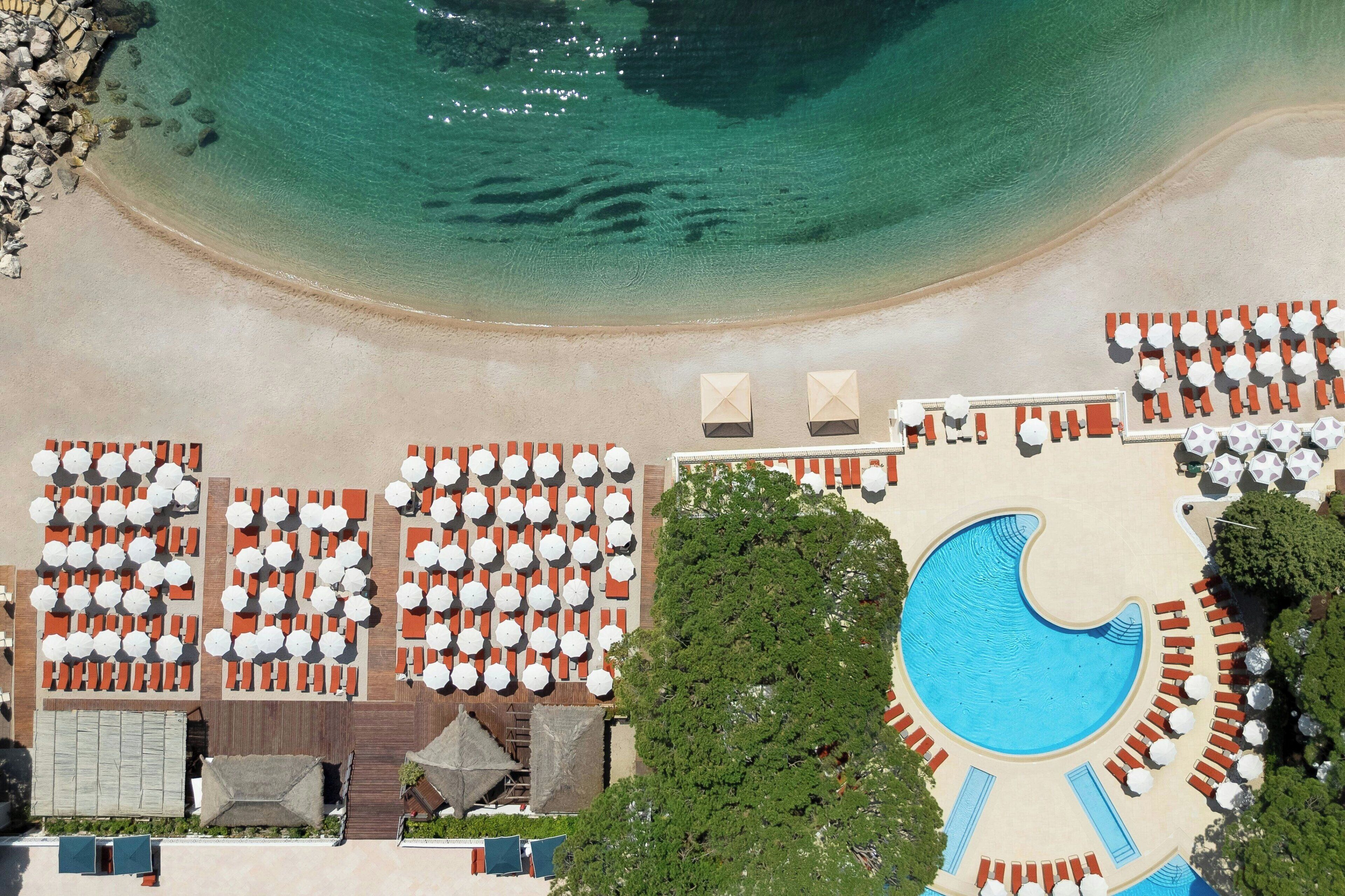 Vue aérienne d’un complexe hôtelier de bord de mer, avec une grande piscine, plusieurs espaces de détente et une plage de sable.