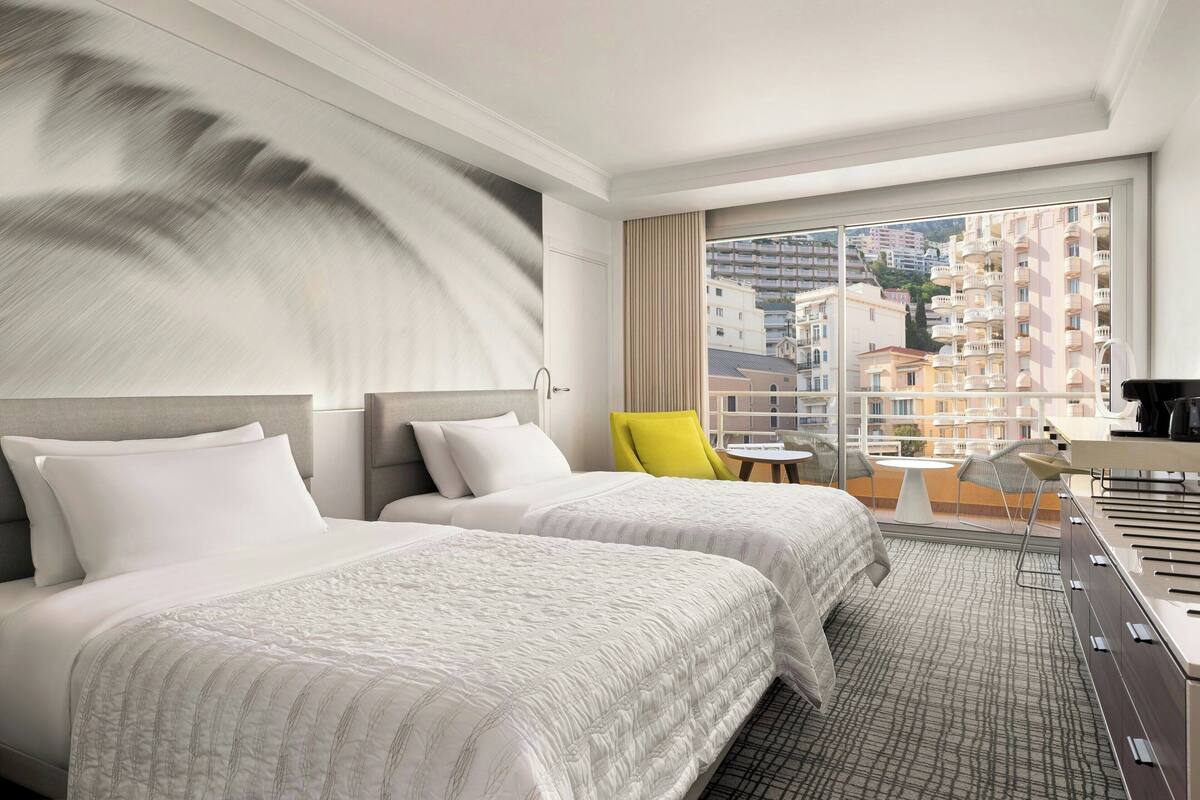 A hotel room with two beds, a large window with a city view, a small table, and a chair.