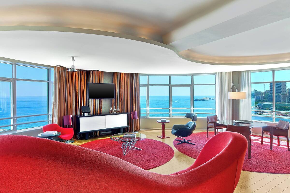 A modern hotel room with a curved ceiling, large windows offering a view of the sea, a flat-screen TV, and red upholstered chairs.