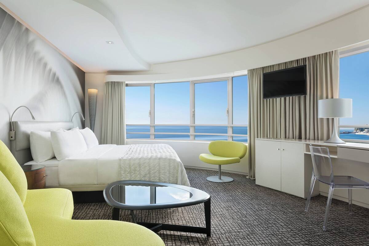 A hotel room with a bed, a desk, a chair, a TV, and a view of the sea.