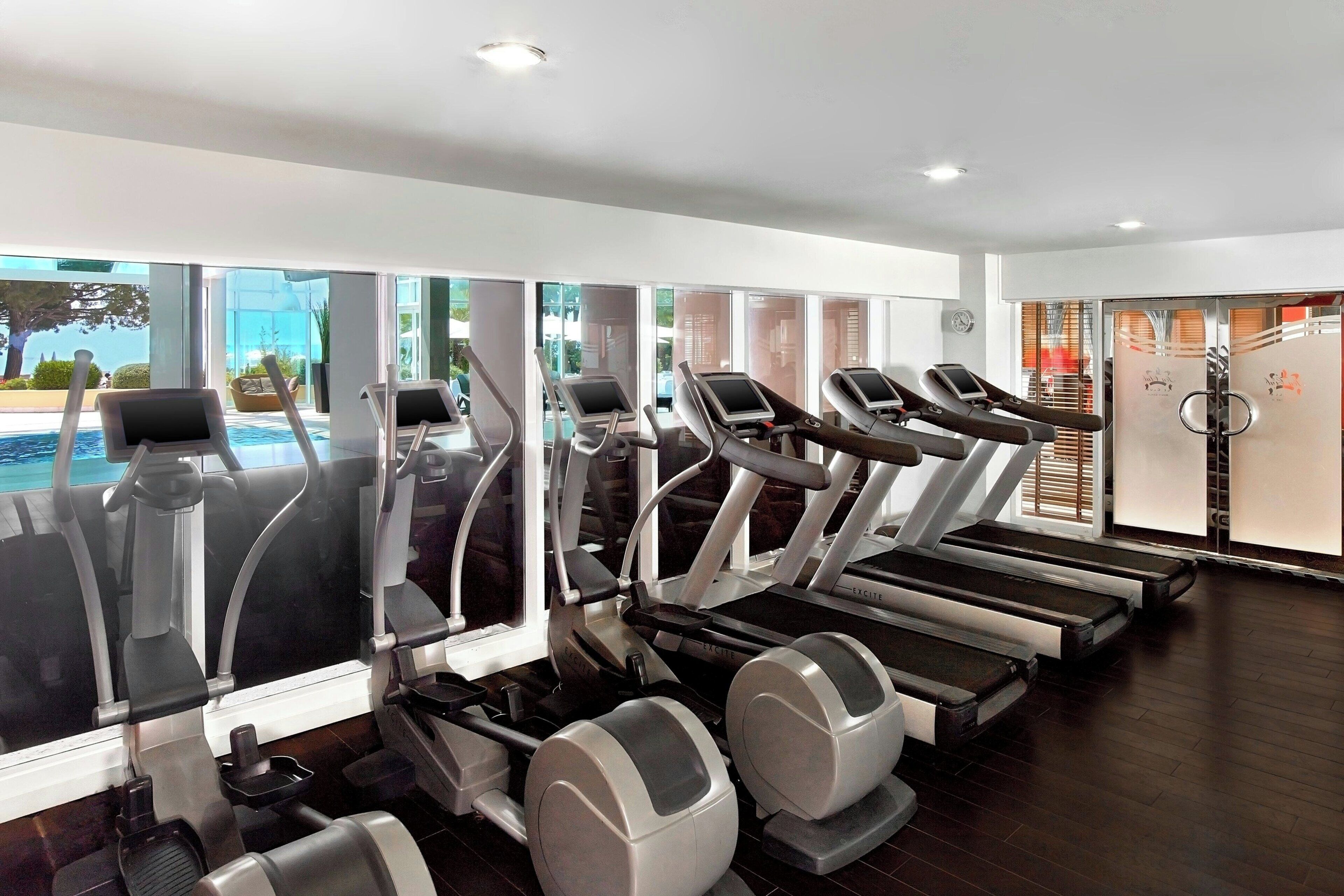 Un gimnasio moderno con cintas de correr, elípticas y una zona de piscina visible a través de amplios ventanales.