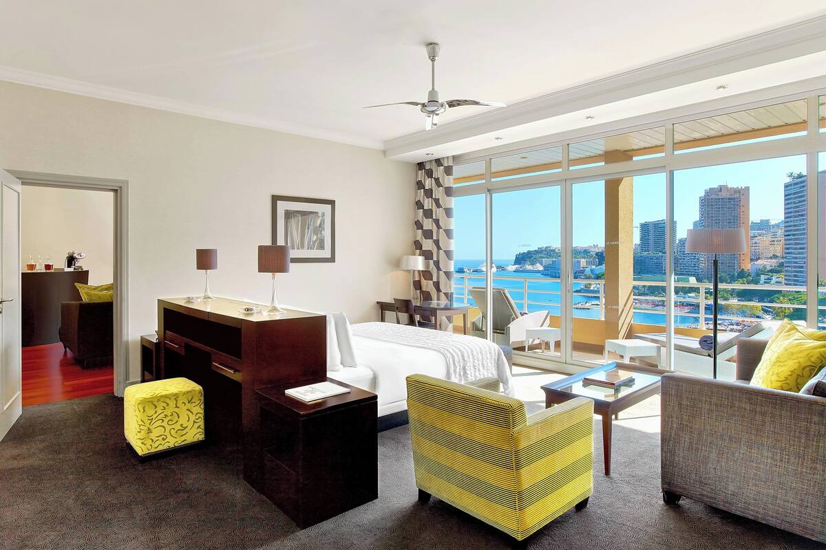 A hotel room with a bed, a desk, a chair, and a sofa. There is a large window with a view of the city and the sea.