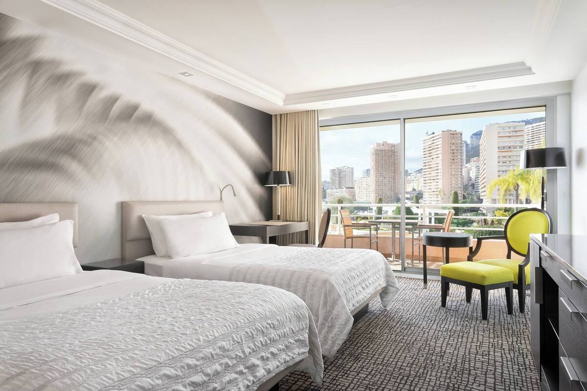 A hotel room with two beds, a desk, a chair, and a balcony with a city view.