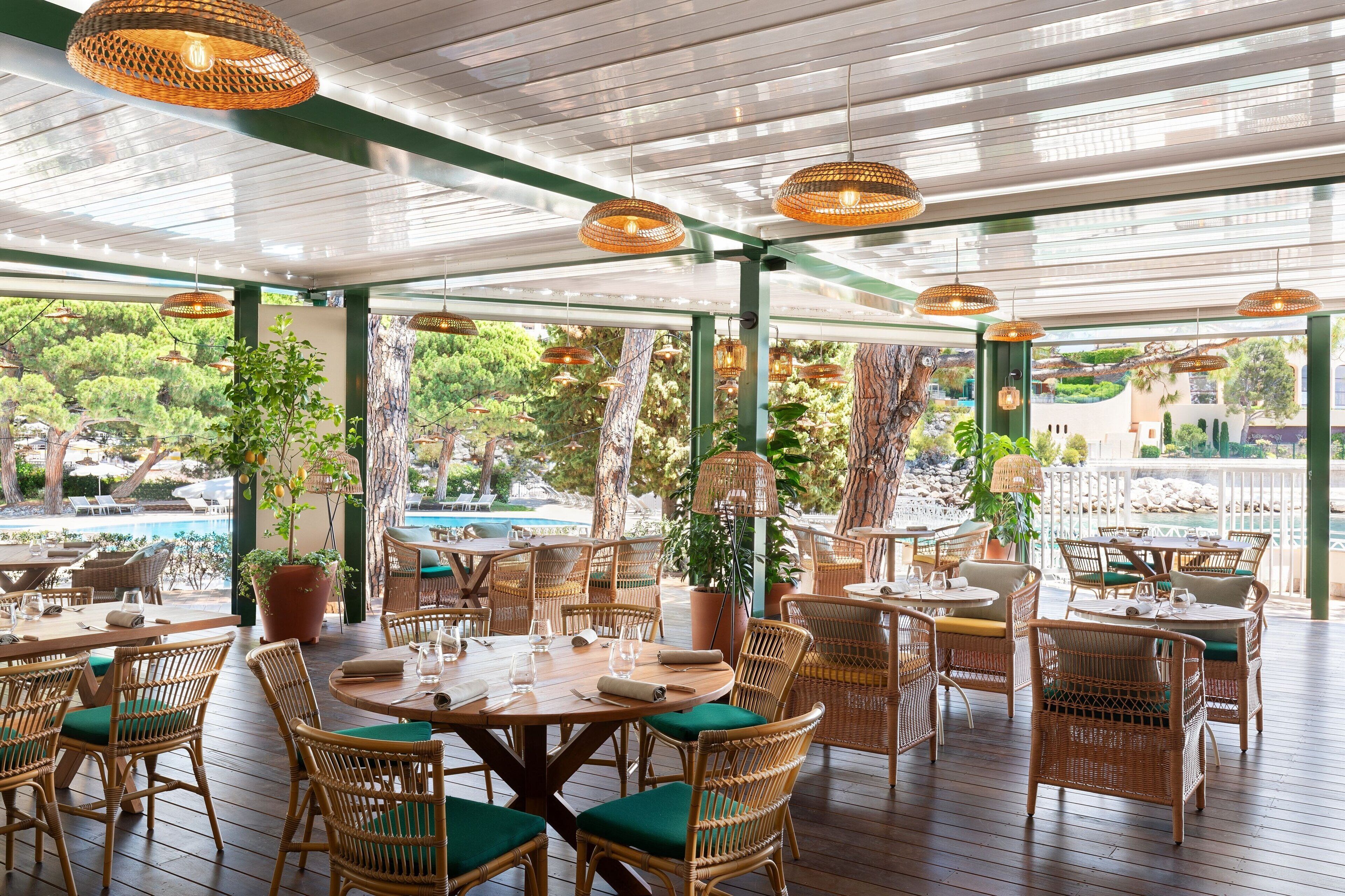 An outdoor dining area with wicker furniture and hanging lights, overlooking a pool and lush greenery.