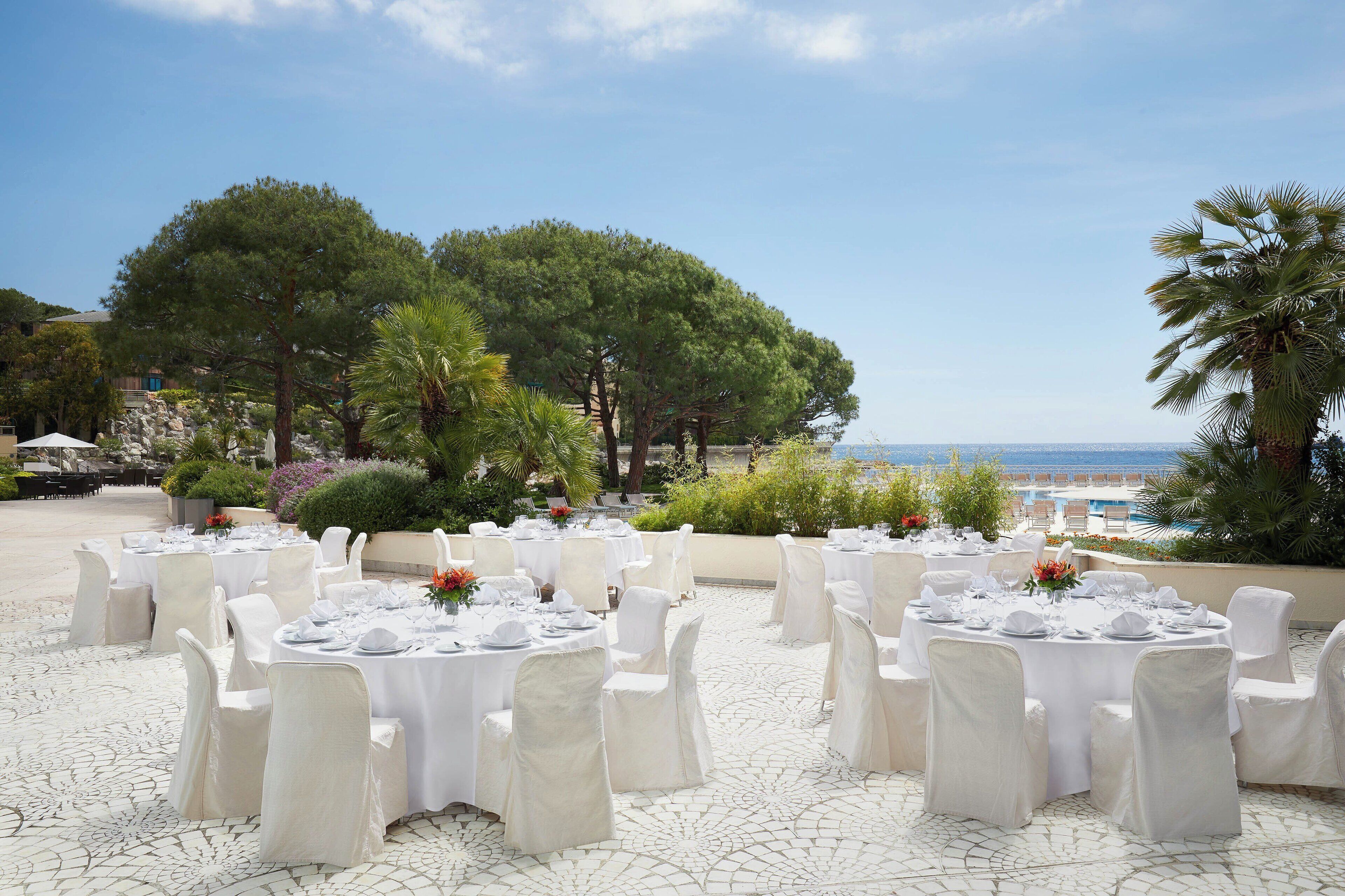 Un área de comedor al aire libre con mesas redondas dispuestas para una comida, rodeada de sillas blancas, y vista al mar y árboles al fondo.