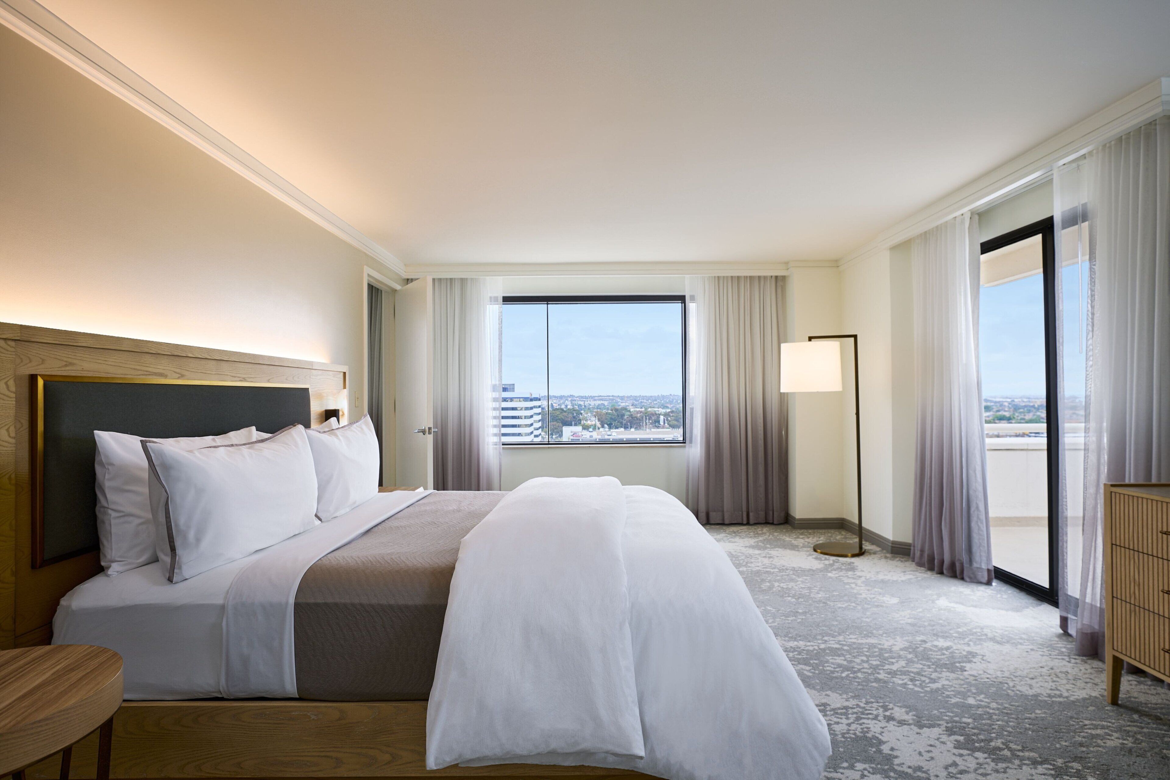 Habitación de hotel con una cama grande, una mesita de noche, una lámpara de pie y vistas a la ciudad a través de la ventana.