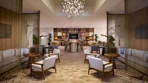 A spacious living area with a central coffee table, sofas, and chairs arranged around it. A television is mounted on the wall, and there are plants and decorative items on shelves.