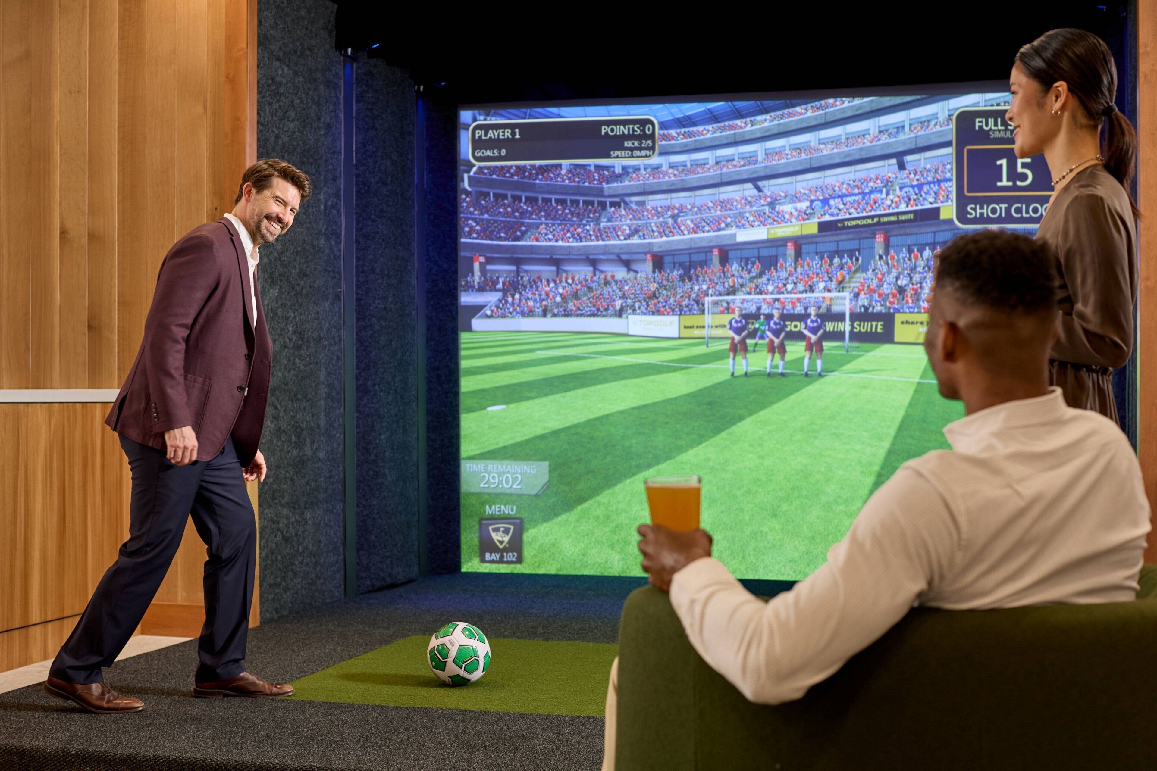 A man in a suit standing on a stage with a soccer ball, a large screen displaying a soccer game, and another person holding a tablet.