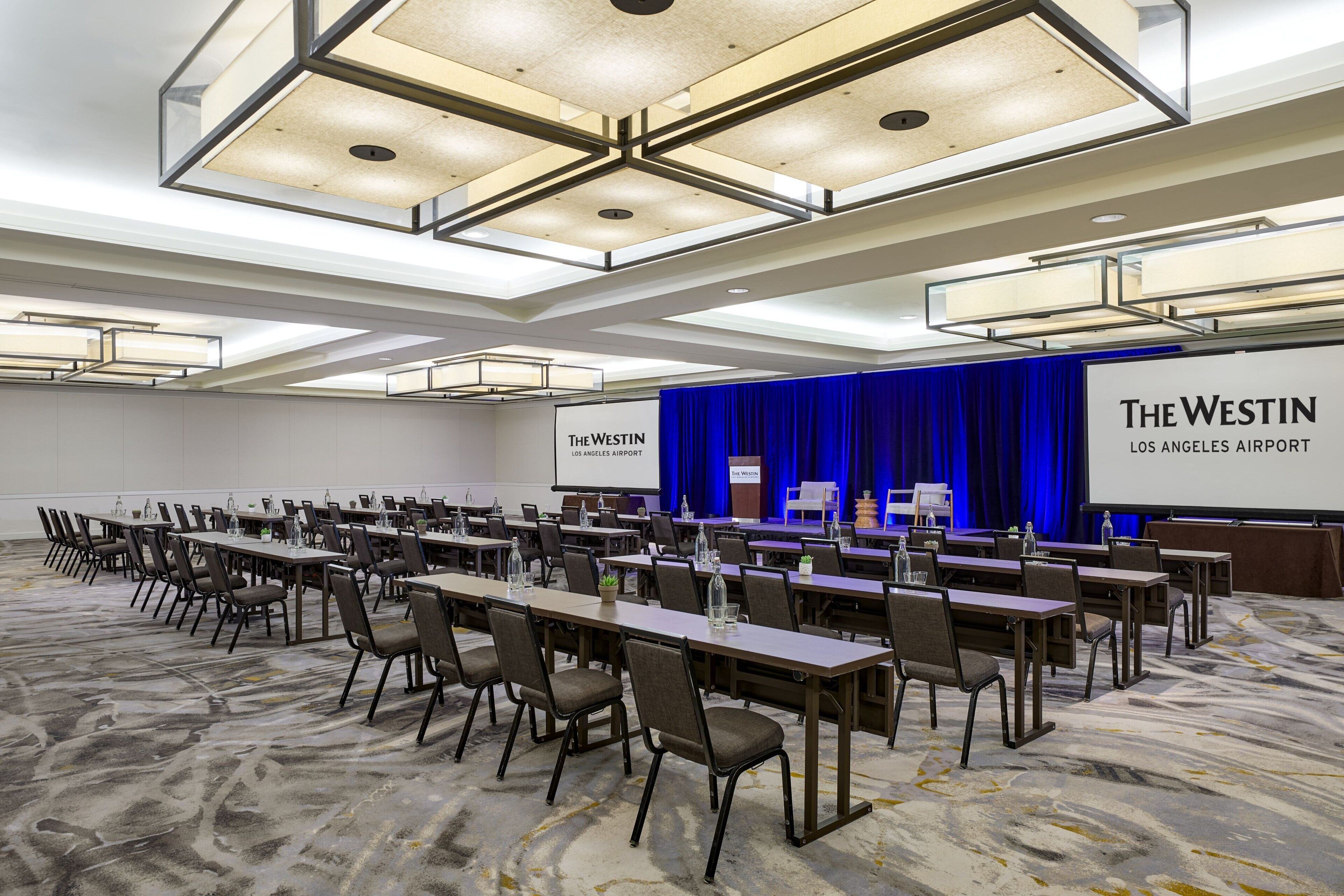 Un salón de conferencias con mesas largas y sillas, un escenario con un telón azul y una pantalla mostrando 'The Westin Los Angeles Airport'.