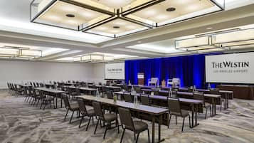 Un salón de conferencias con mesas largas y sillas, un escenario con un telón azul y una pantalla mostrando 'The Westin Los Angeles Airport'.