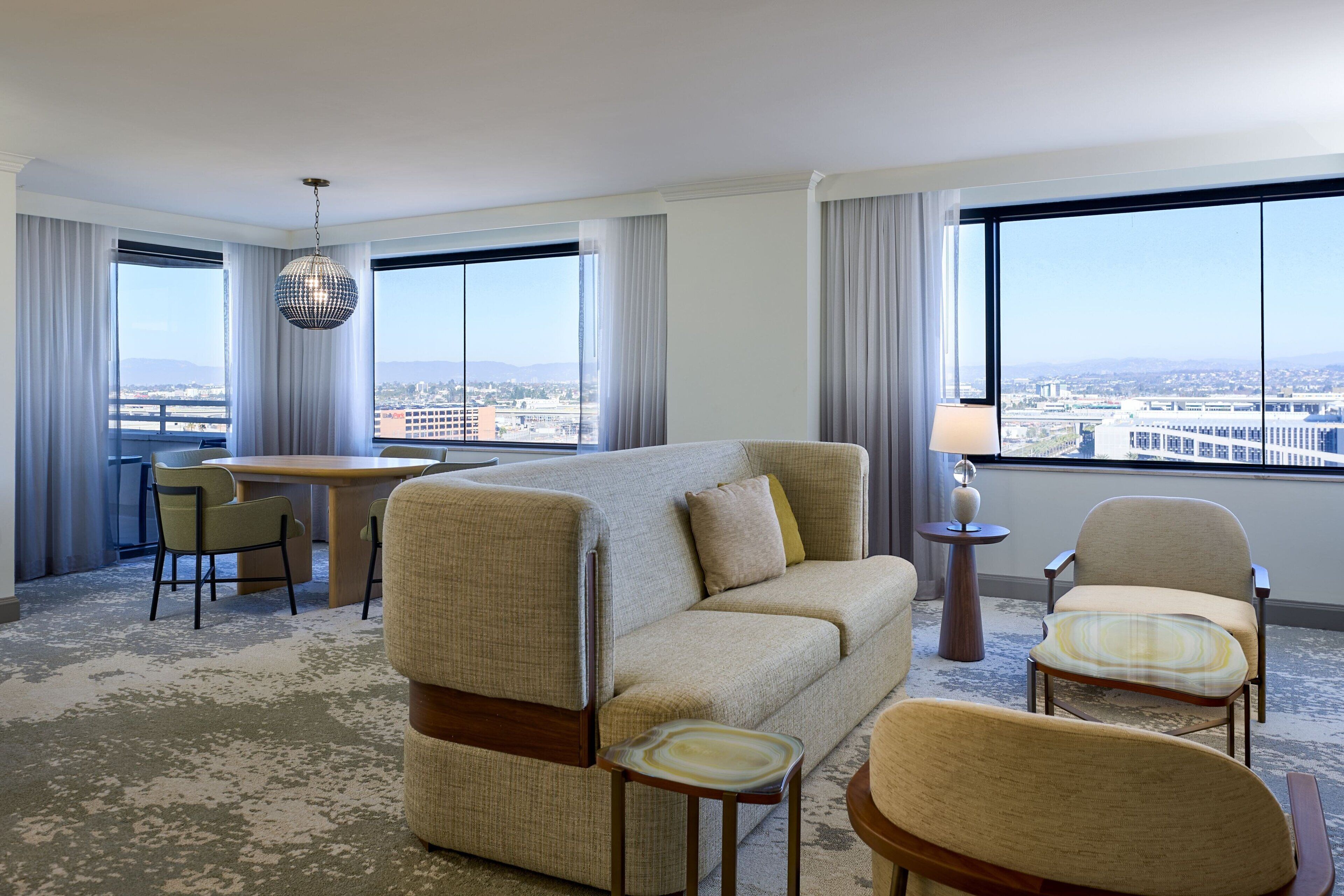 Una camera d'hotel con divano, sedie, un tavolo da pranzo e vista sulla città attraverso ampie finestre.