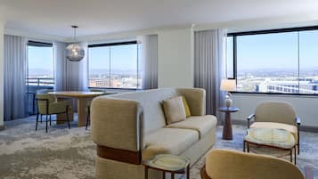A hotel room with a sofa, chairs, a dining table, and a view of a cityscape through large windows.