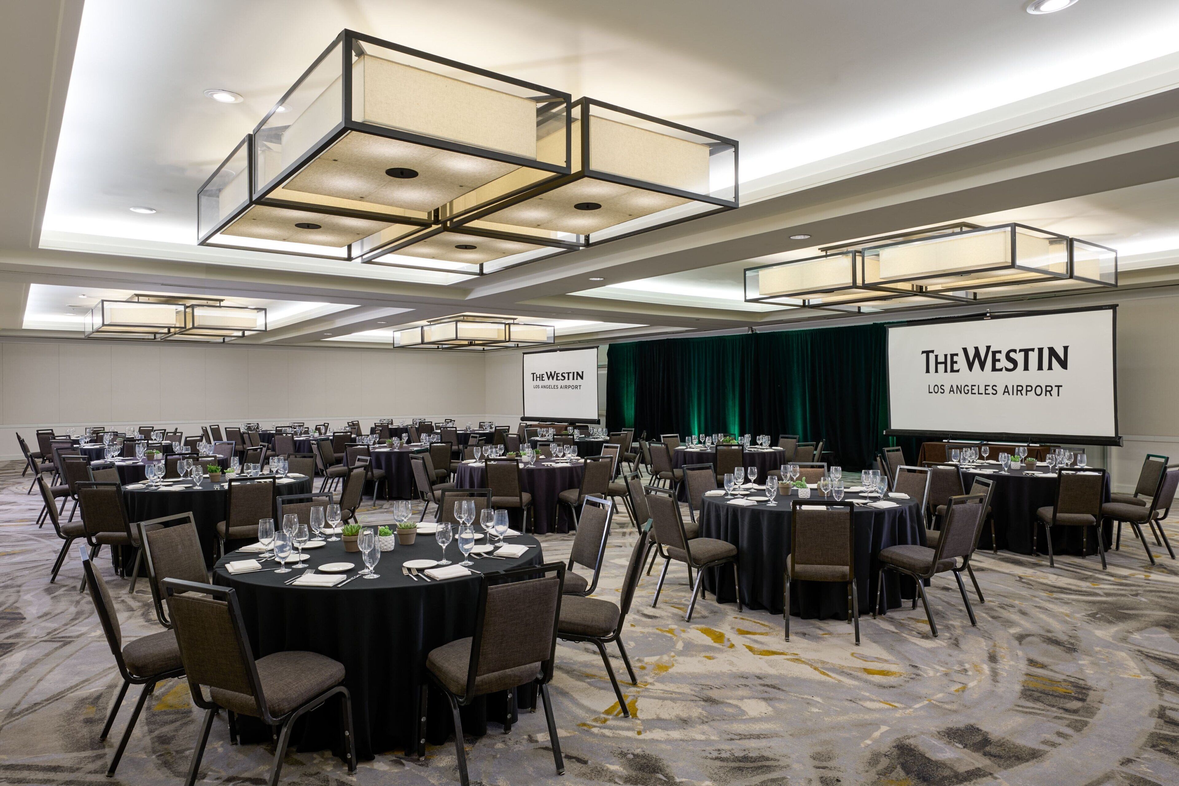 Ein Konferenzraum mit runden Tischen, Stühlen und einem großen Bildschirm, der 'The Westin Los Angeles Airport' anzeigt.