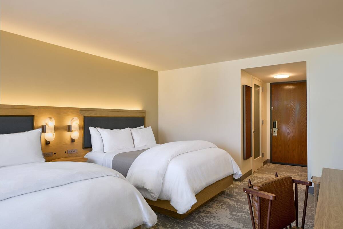 Habitación de hotel con dos camas, cabecero de madera, lámparas fijadas a la pared, puerta de madera y una pequeña silla de madera.