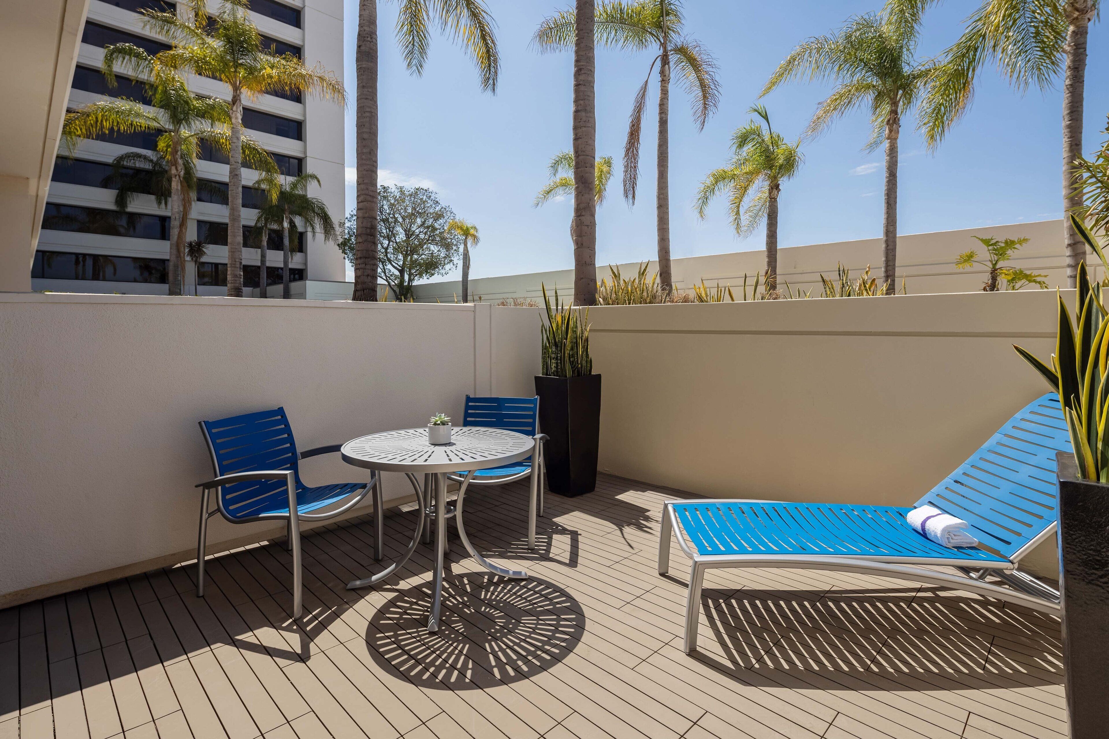 Una terraza en la azotea con una mesa y sillas, un sillón de descanso y altas palmeras.