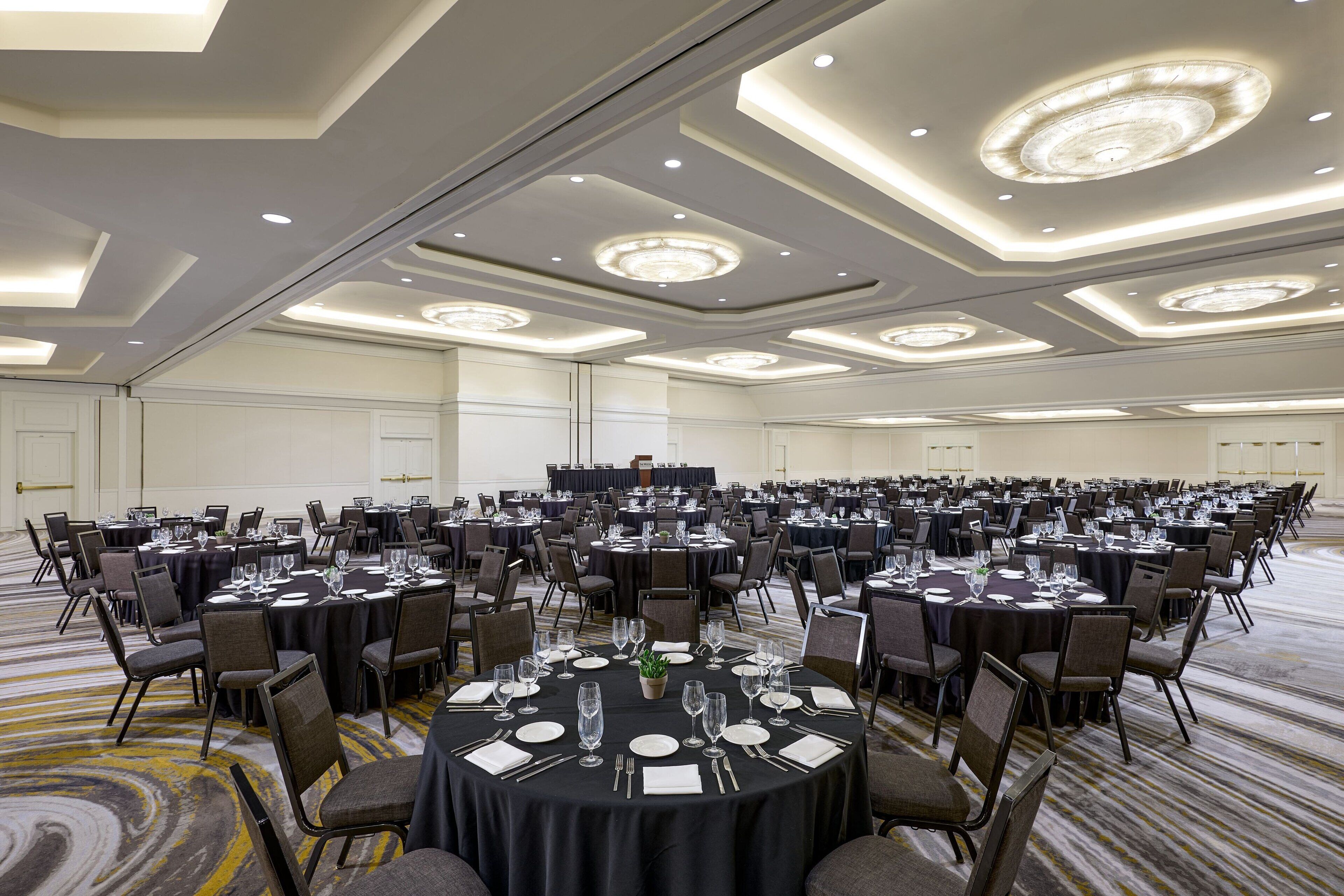 Une grande salle de conférence équipée de plusieurs tables rondes, chacune agrémentée de chaises, d’assiettes, de couverts et de verres à vin.