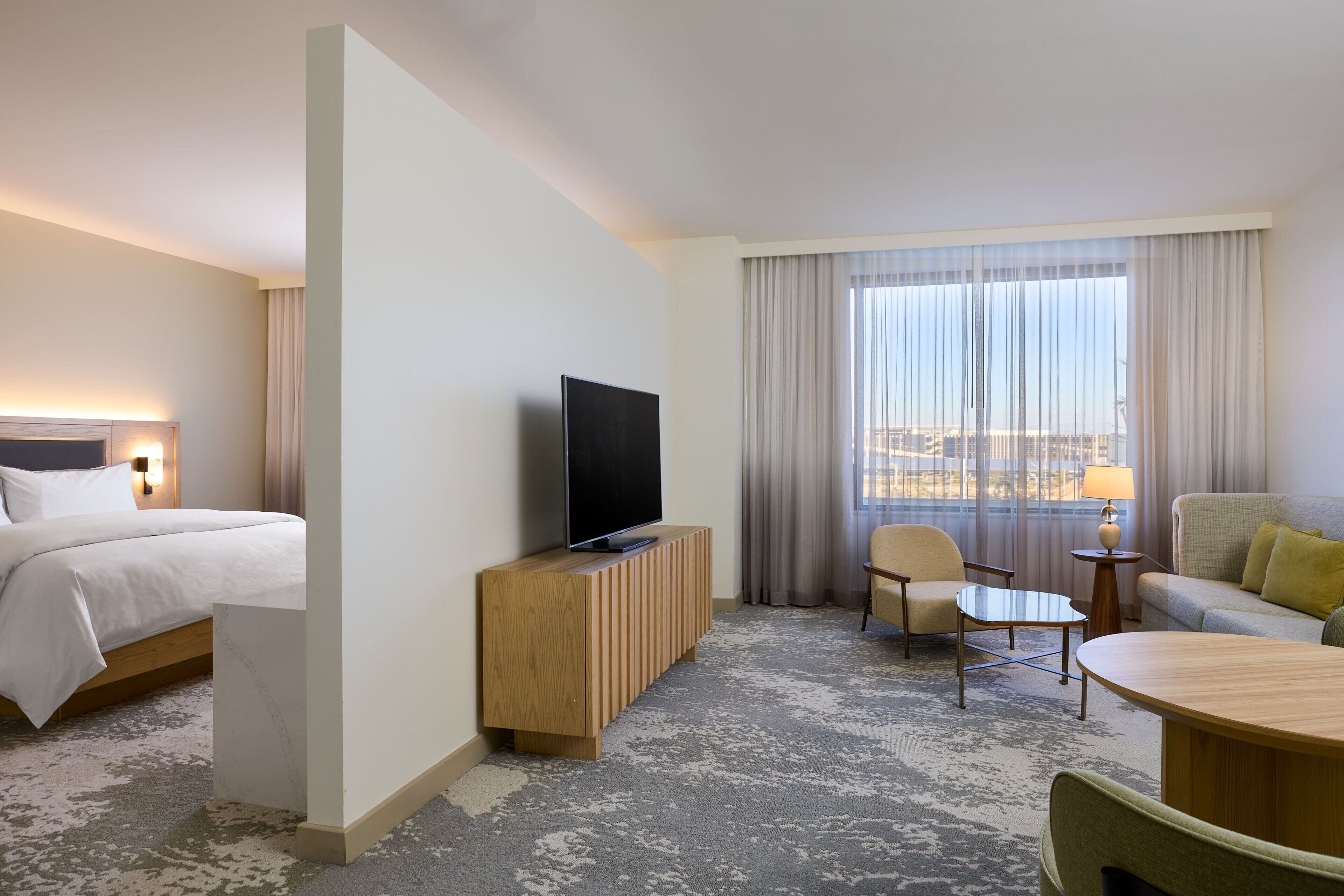 Ett hotellrum med en säng, en tv-apparat placerad på en träkonsol, en sittgrupp med ett bord och stolar samt ett stort fönster med gardiner.
