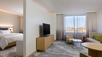 Une chambre d’hôtel comprenant un lit, un téléviseur posé sur une console en bois, un coin salon avec une table et des chaises, et une grande fenêtre avec des rideaux.