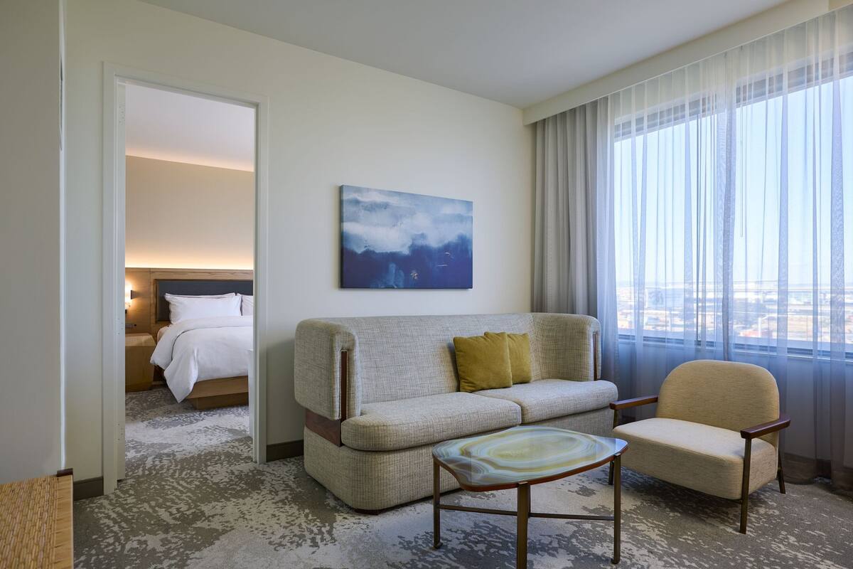 Habitación de hotel con sofá, sillón y una mesita. Se observa un cuadro en la pared y una ventana con cortinas.