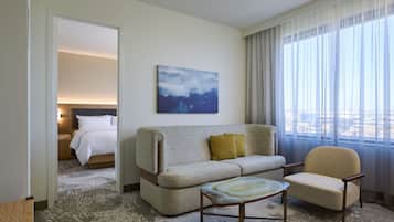 A hotel room with a sofa, armchair, and a small table. There is a painting on the wall and a view of the city through the window.