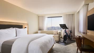 A hotel room with a large bed, a sofa, a desk with a chair, a television, and a view of the city through the window.