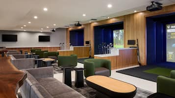 Et moderne loungeområde med flere siddepladser, et billardbord og et projektorskærm, der viser en golfkamp.