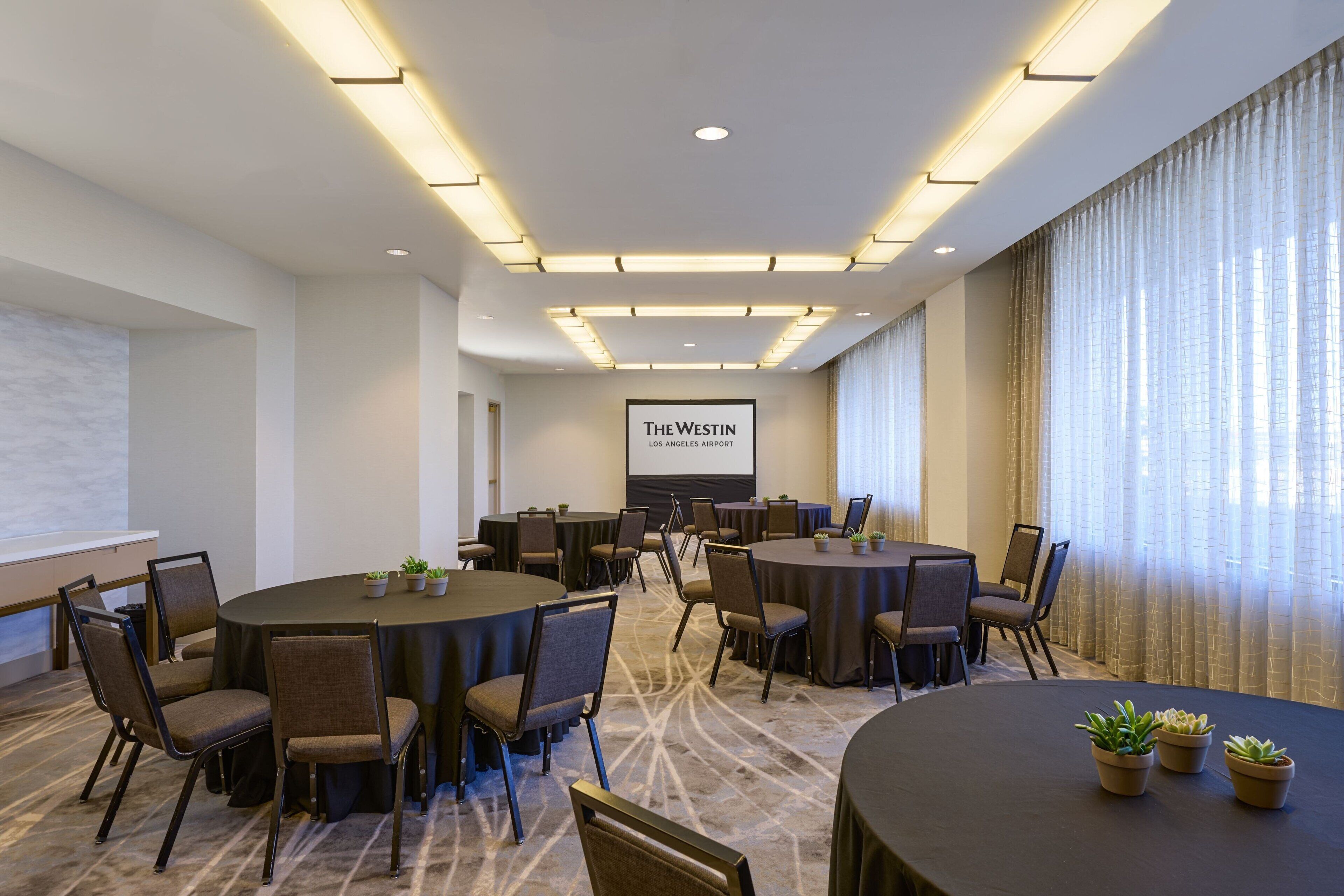 Una sala de conferències amb diverses taules i cadires, una pantalla gran i un pancarta amb la inscripció 'The Westin Los Angeles Airport'.