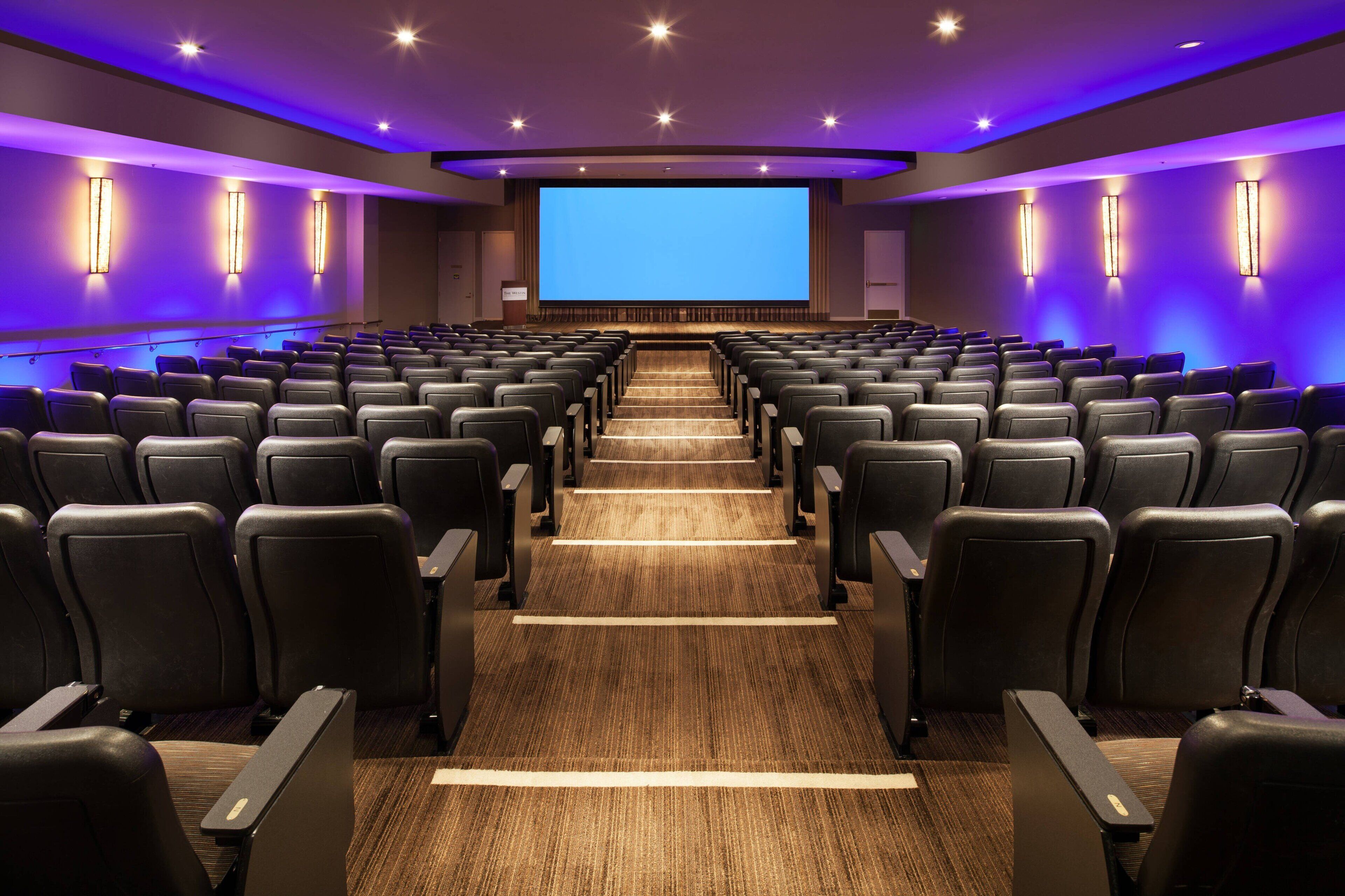 A theater with rows of black seats, a large screen, and purple ambient lighting.