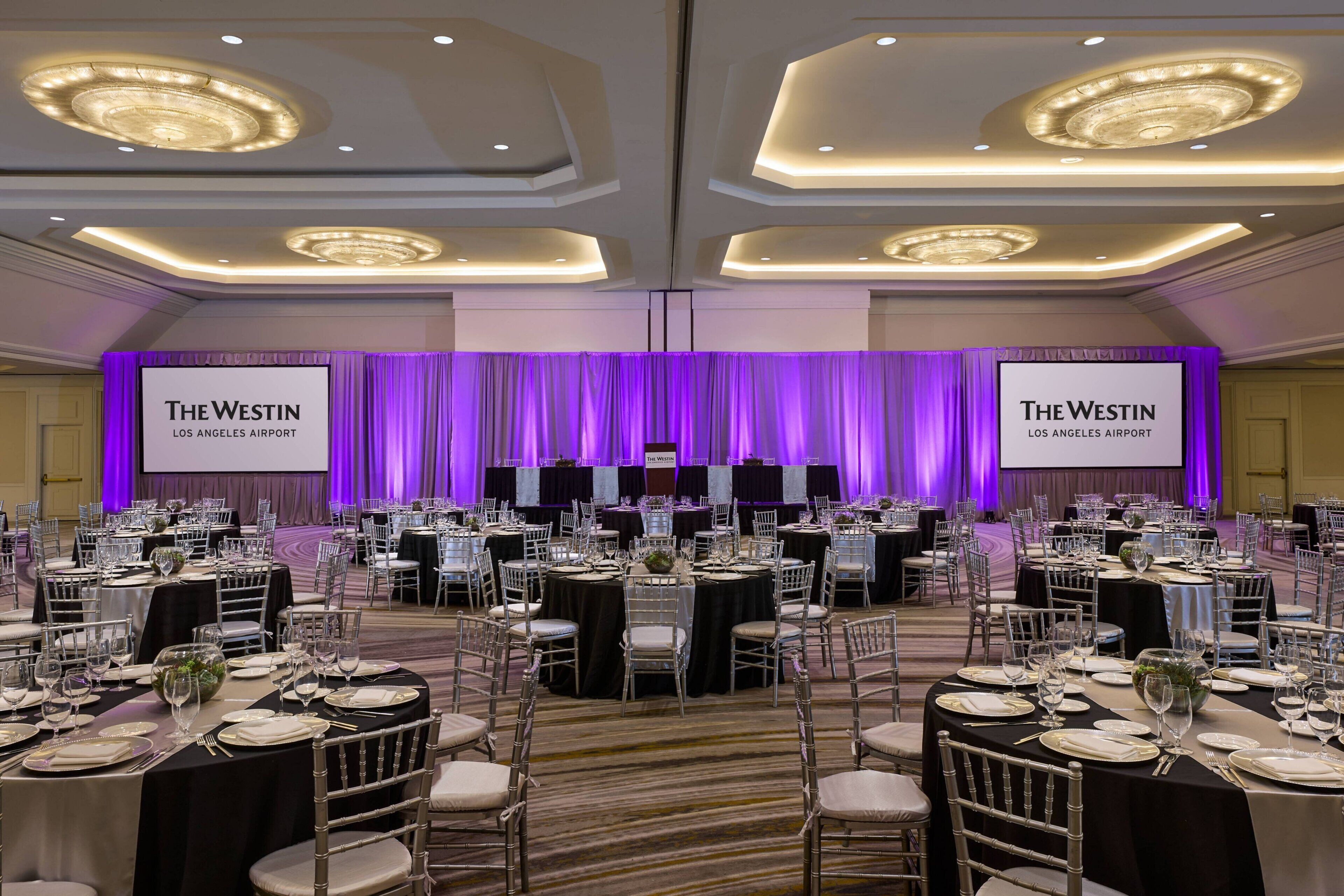 A conference room at The Westin Los Angeles Airport with multiple round tables, chairs, and a stage with a purple backdrop.