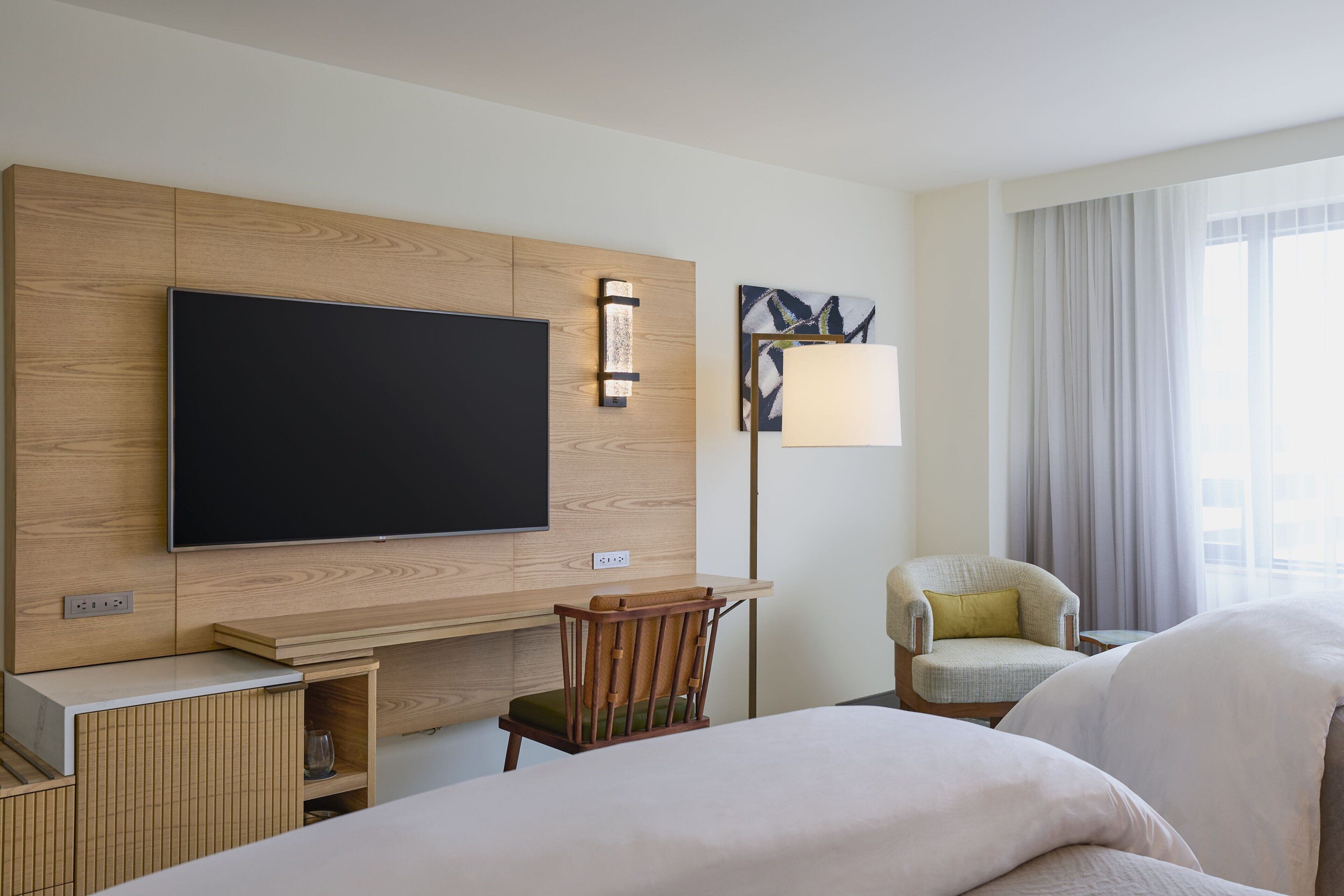 A hotel room with a flat-screen TV, a desk with a chair, a lamp, and a bed.