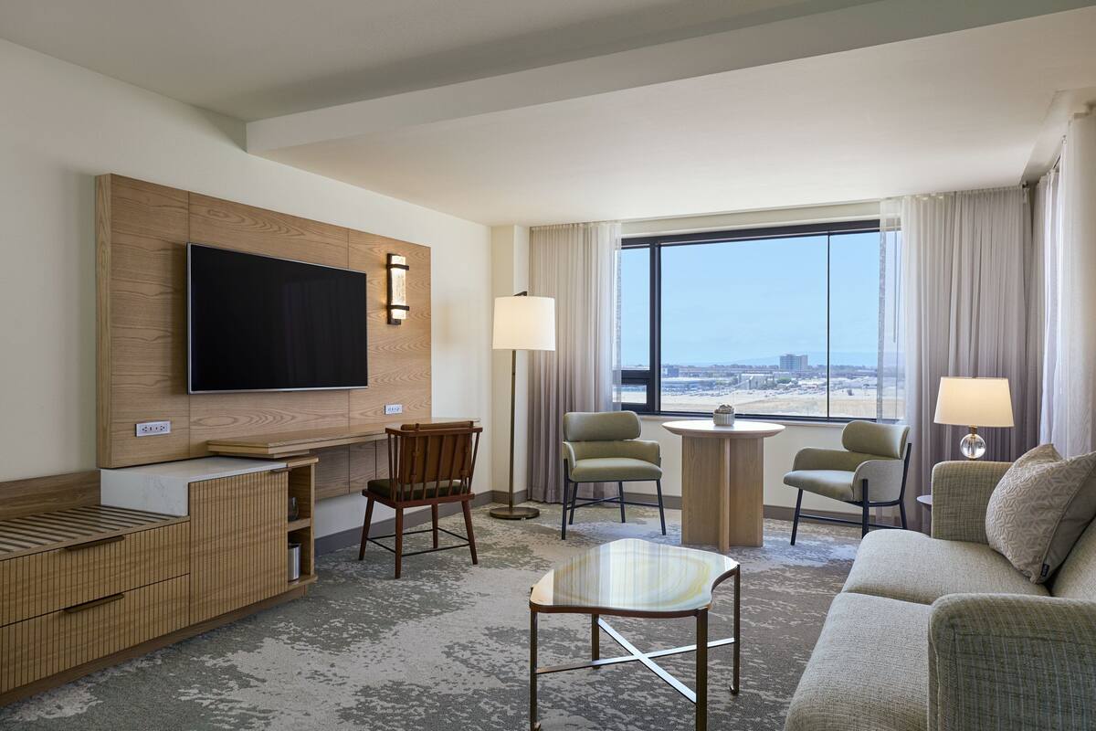Habitación de hotel con un televisor de pantalla plana montado en un panel de madera, un sofá, dos sillones, una mesita y vistas a la ciudad a través de amplios ventanales.