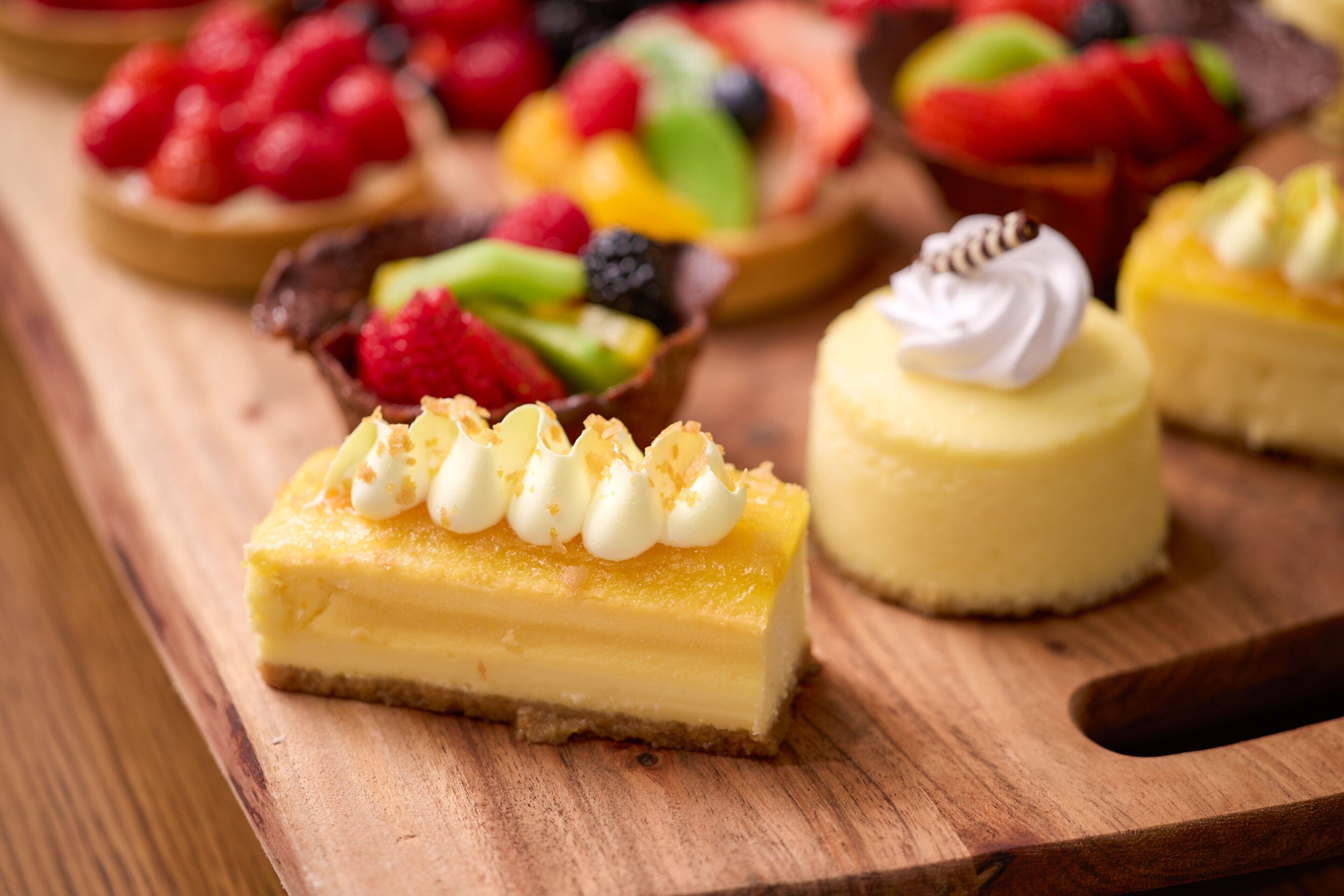 A slice of cheesecake with whipped cream and a fruit-topped tart on a wooden board.