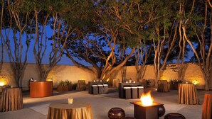 Un área de comedor al aire libre con mesas redondas, una chimenea central y un fondo de árboles y un muro de piedra.