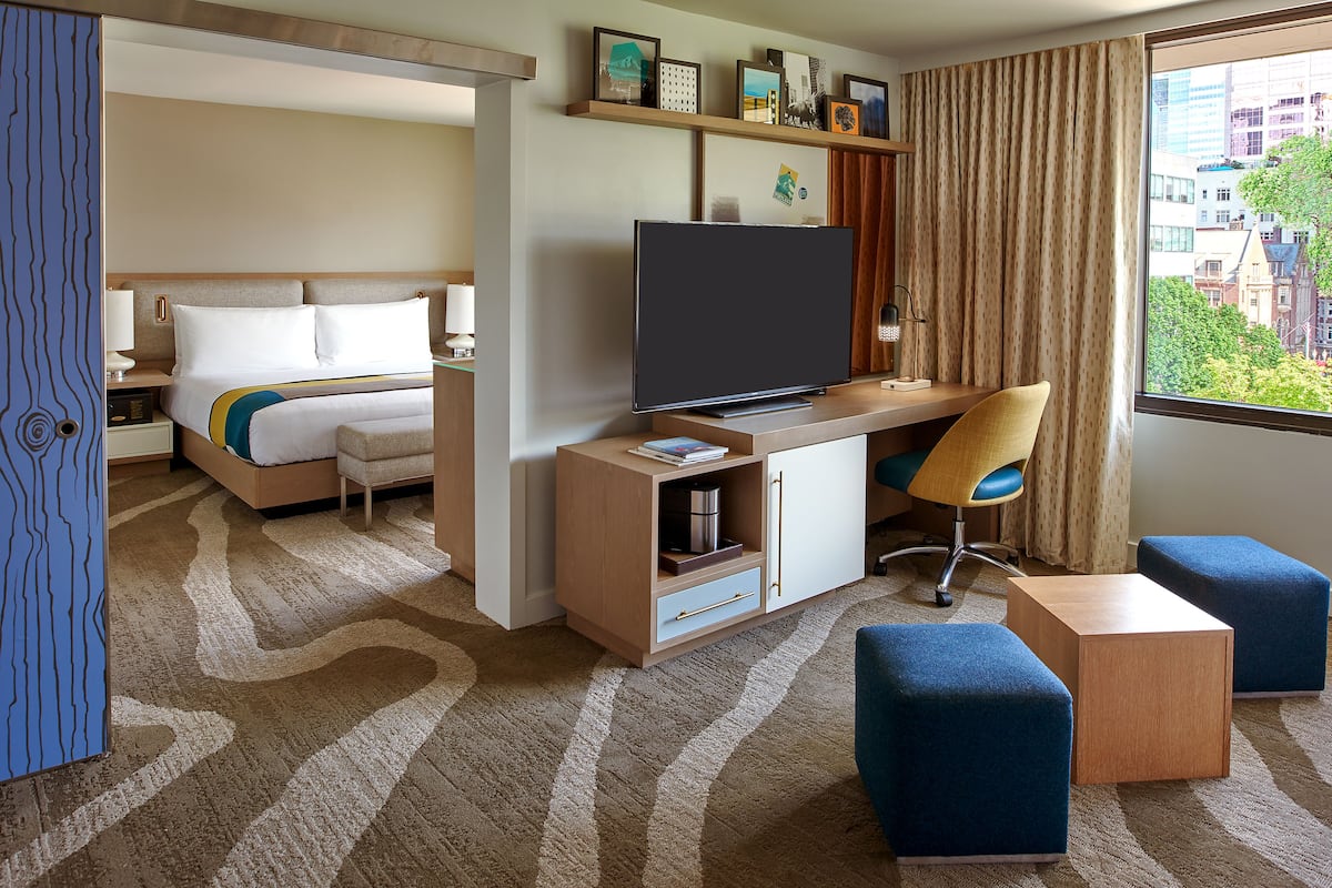 A hotel room with a bed, a desk with a TV, a chair, and two ottomans.