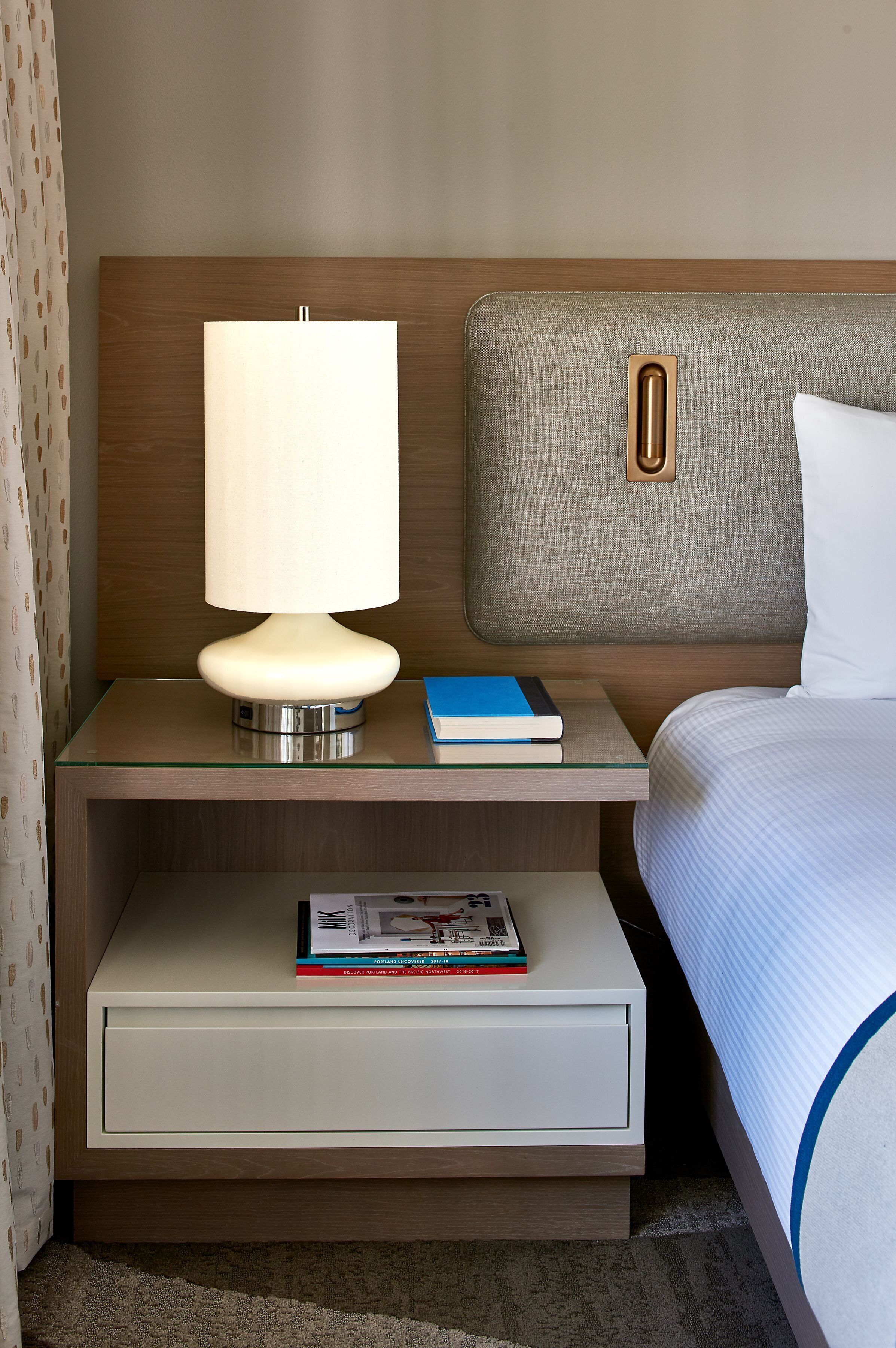 A neatly arranged hotel room with a bed, a bedside table with a lamp, and a nightstand with a book and a magazine.