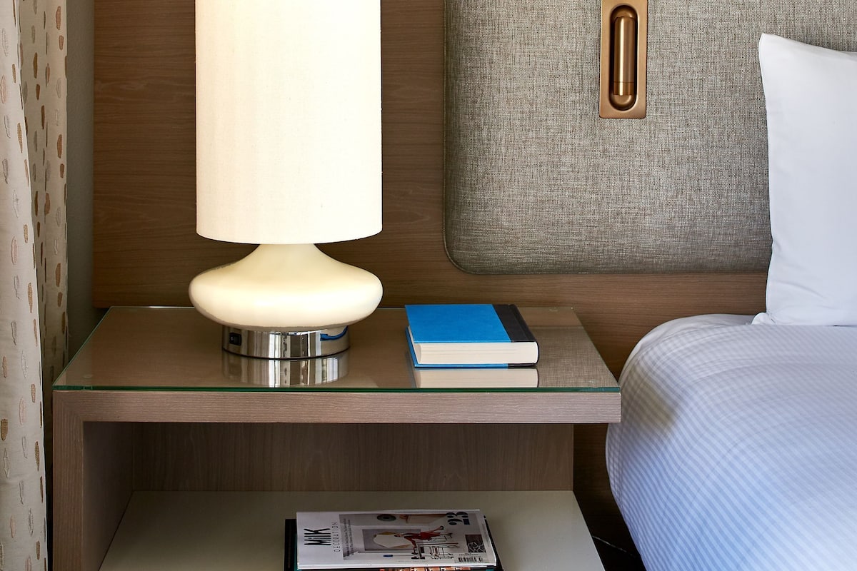 A neatly arranged hotel room with a bed, a bedside table with a lamp, and a nightstand with a book and a magazine.